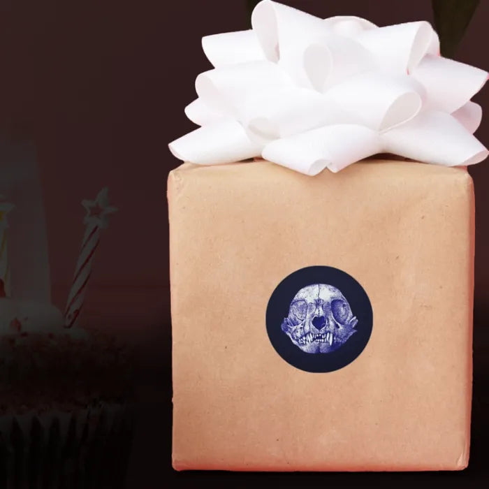 Sticker of cat skull drawing tinted purple on a black circle, prototyped on the side of a wrapped gift.