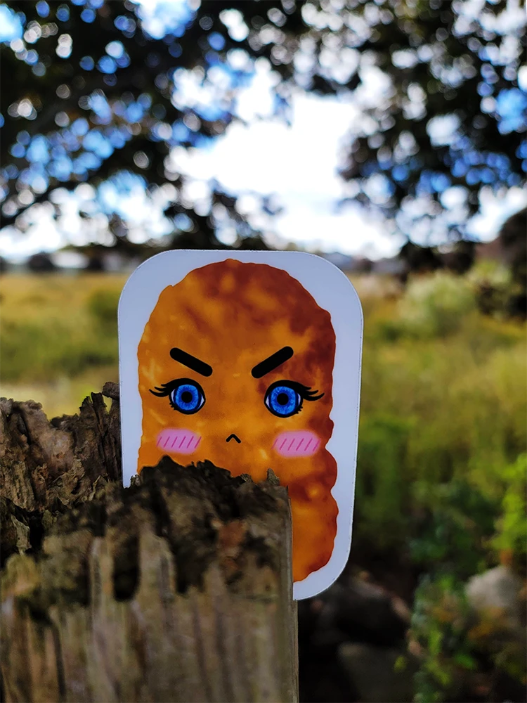 Grumpy chicken nugget sticker is wedged into the top of a wooden post. Behind it is a green field and blue sunny sky with tree branches making shade.