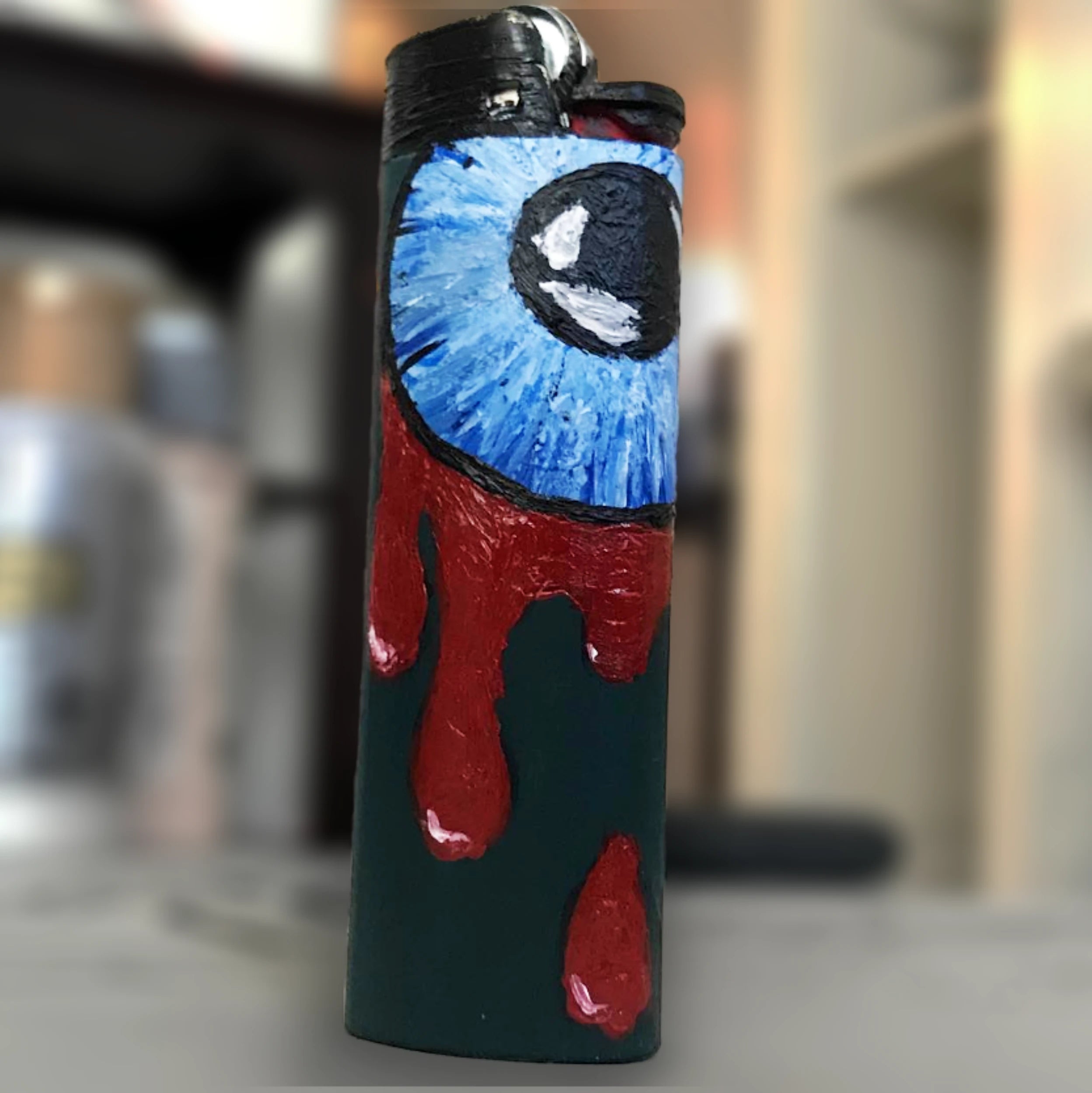 Painting on a bic lighter of a bright blue circular eyeball with dripping blood.
