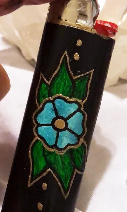 Painting on a bic lighter of a small blue flower with green leaves in traditional tattoo style. 
