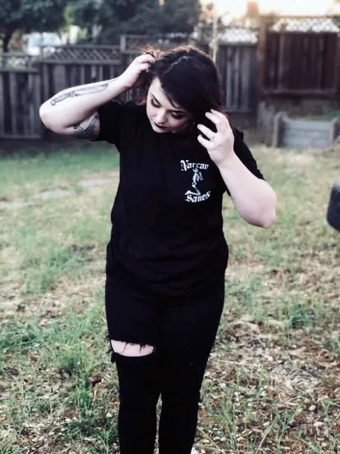 A woman is wearing Narcan Saves shirt. She is looking down, facing the viewer, standing in a  backyard. 