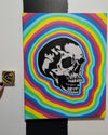 painting of skull with a rainbow background hanging  on a black and white striped wall. a small sticker version is held up next to it.