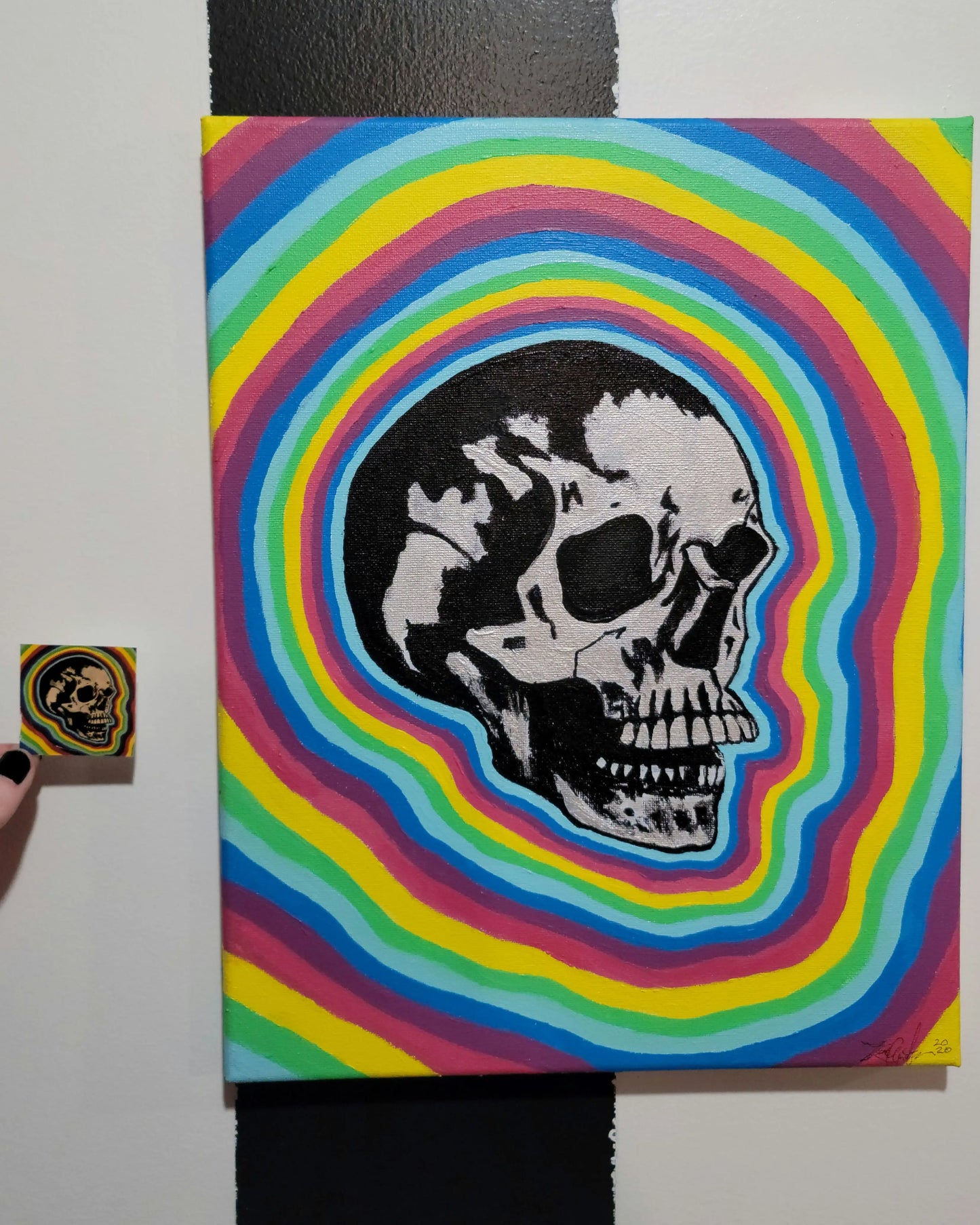 painting of skull with a rainbow background hanging  on a black and white striped wall. a small sticker version is held up next to it.