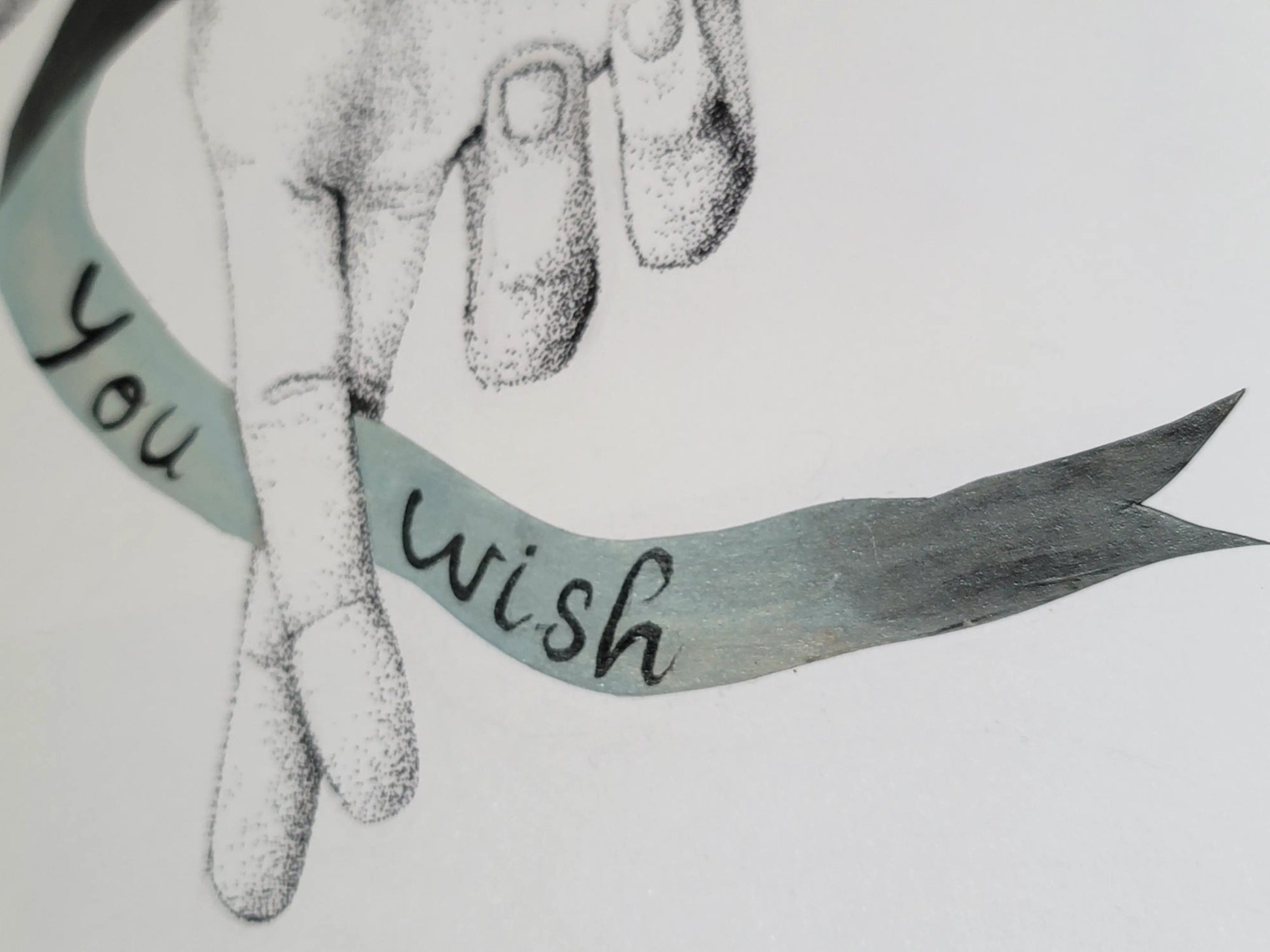 close up of "as you wish" at a slight side angle. it shows the two crossed fingers and part of the other two folded over fingers. the blue-gray ribbon wraps in from the left and goes between the crossed fingers. on the left side the ribbon says "you" and on the right it says "wish".