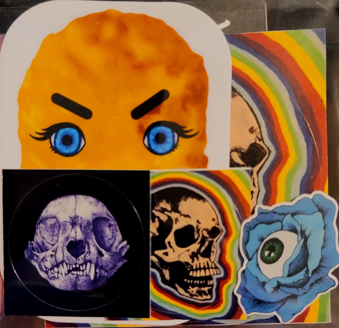 a clear envelope showing the contents of a sticker pack. the top half this item can be seen in the top left, along with a few other stickers like a purple cat skull, rainbow skull  stickers, and a blue rose with an eyeball. 